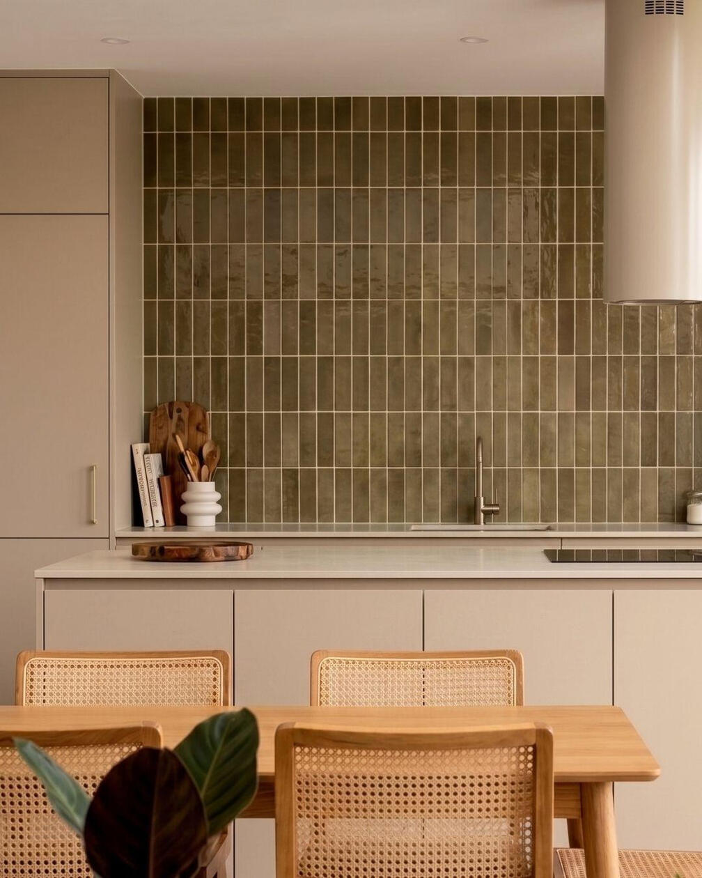 Uma paleta de materiais naturais ganha profundidade através do contraste entre madeira, pedra e azulejo. Cozinha com armários claros, bancada em madeira, parede revestida a azulejo verde e zona de refeições com mesa e cadeiras em madeira.