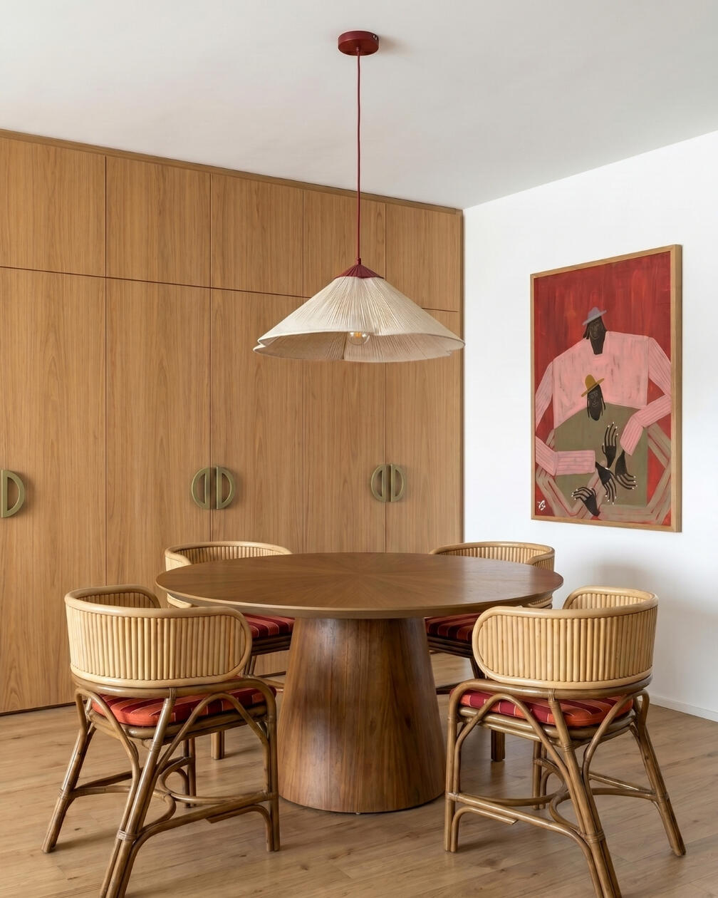 Sala de jantar com mesa redonda em madeira, quatro cadeiras em fibras naturais, candeeiro suspenso e parede revestida a madeira.