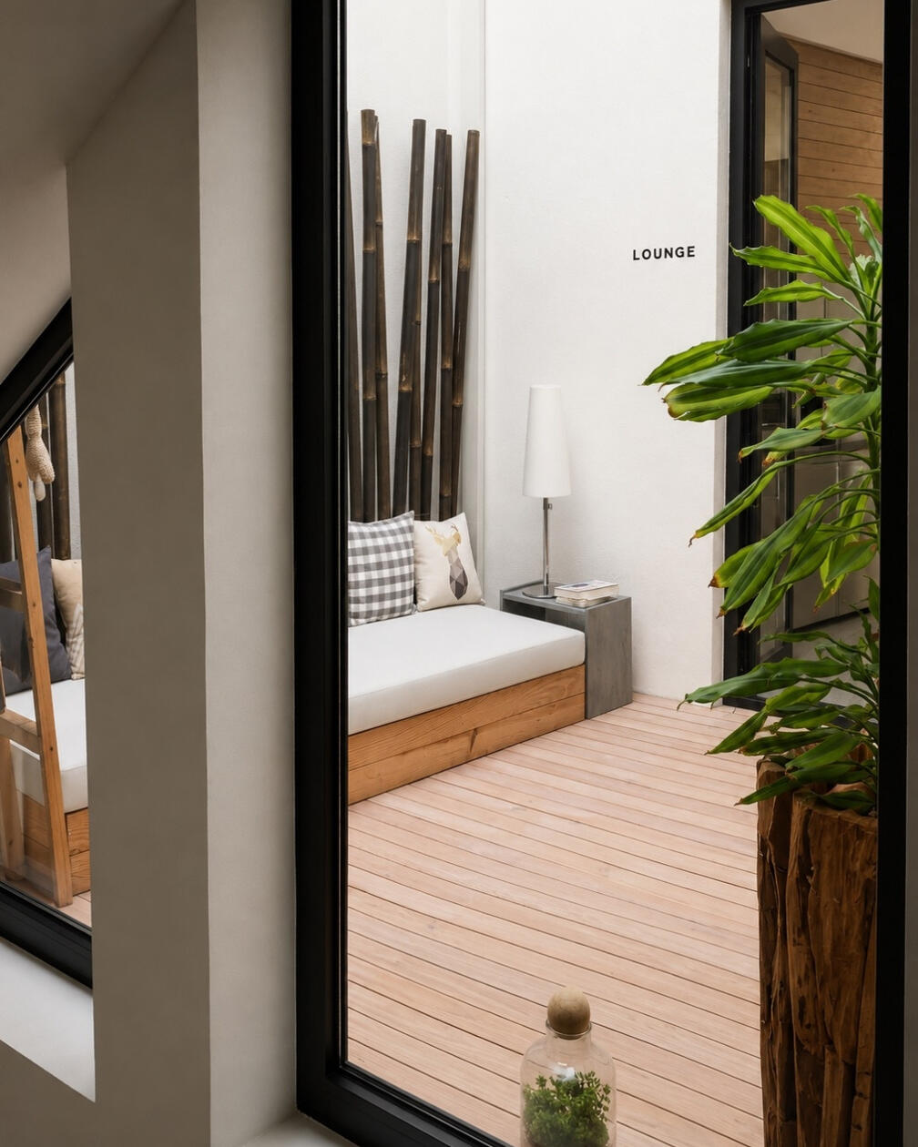 Um pequeno lounge exterior onde madeira, vegetação e materiais naturais prolongam a experiência da casa. Lounge exterior com banco em madeira, almofadas neutras, mesa lateral, candeeiro e vegetação junto a uma parede branca com elementos em bambu.